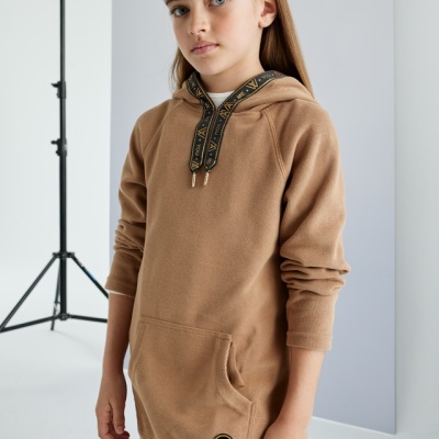 Criança com hoodie castanho e cordões pretos com letras douradas num estúdio minimalista