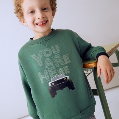 Criança com camisola verde com impressão 'YOU ARE HERE' e carro preto