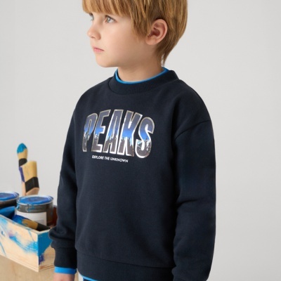 Menino loiro a vestir sweatshirt preta com texto PEAKS e EXPLORE THE UNKNOWN, fundo cinzento, caixa com tinta azul