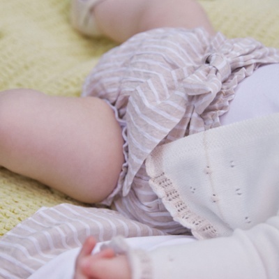 Roupa de bebé com calções às riscas e body branco, sapatinhos rosa claro, sobre manta amarela tricotada
