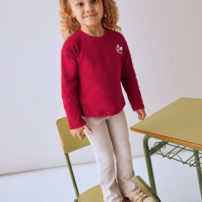 Criança com camisola vermelha, calças brancas e sapatilhas douradas em cadeira escolar verde.