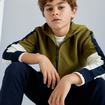 Criança com camisola verde-oliva, branco e azul-marinho, calças azul-marinho, sentada numa superfície de madeira com fundo cinzento