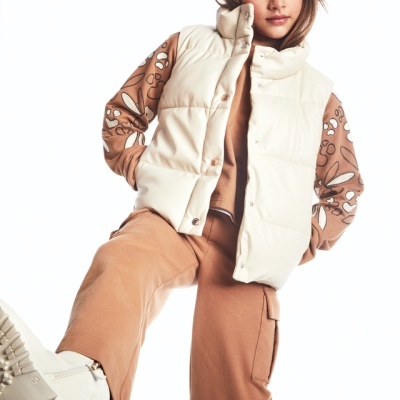 Criança com colete creme, camiseta bege com padrão nas mangas, calças castanhas e botas brancas, fundo branco