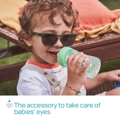 Criança com óculos de sol verdes segurando copo com tampa verde sentada em banco de madeira com almofadas