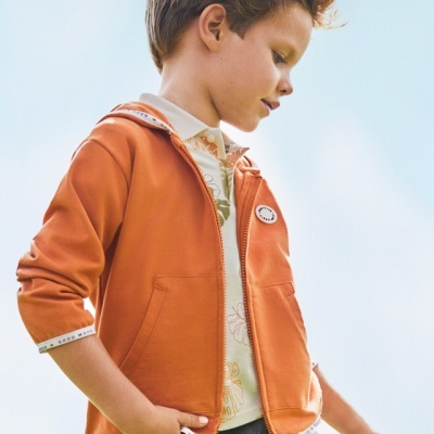 Menino com casaco laranja, camisa branca com padrão e calças cinzentas ao ar livre
