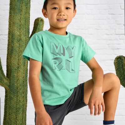 Criança com roupa casual verde e cinzenta, boné e sapatilhas, com cactos e muro branco ao fundo.