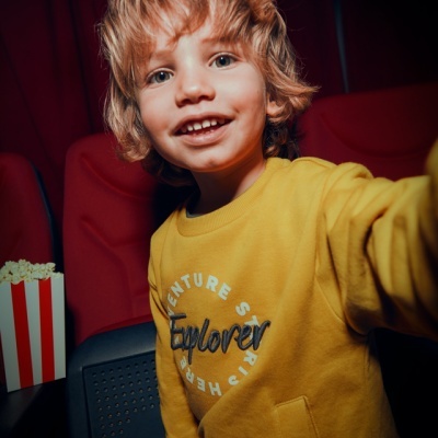 Criança sorridente com camisola amarela em cadeira de cinema vermelha e balde de pipoca