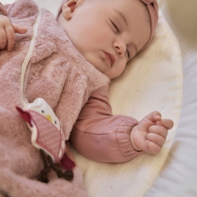 Bebé adormecida de roupa rosa de plush e faixa de cabeça, deitada em fundo branco acolchoado.