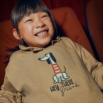 Casaco bege com cão e texto 'HEY THERE friend' usado por criança sentada