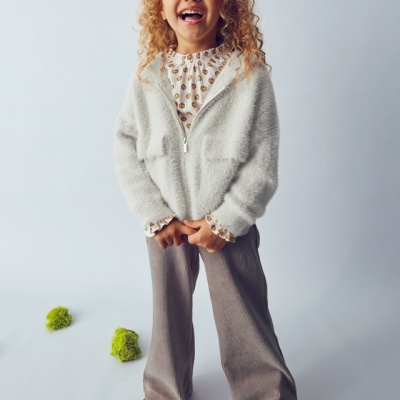 Menina com casaco branco, blusa com padrão castanho, calças cinza largas e botas castanhas em fundo neutro