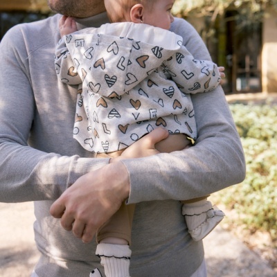Homem com camisola cinza claro segurando bebé com casaco de corações e sapatinhos brancos ao ar livre