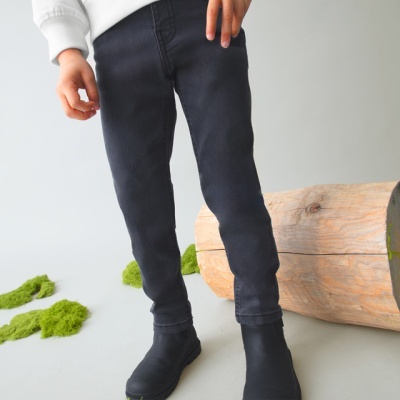 Criança vestindo calças pretas justas e botas pretas, perto de tronco de madeira e musgo verde no chão