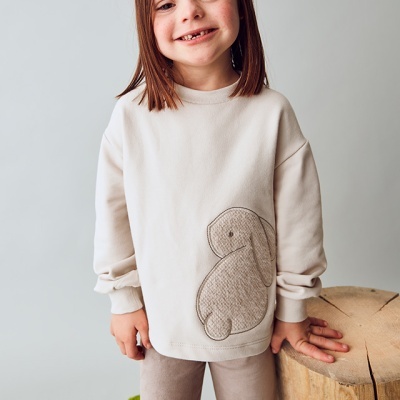 Menina com camisola creme e padrão de coelho apoiada num tronco de madeira