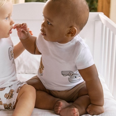 Dois bebés sentados numa cama branca, um vestido com t-shirt branca e calças curtas castanhas.
