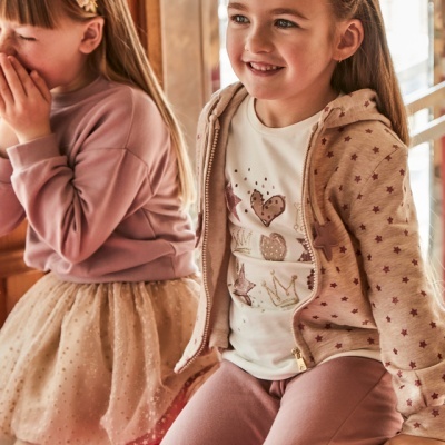 Duas meninas sentadas com roupa rosa e bege, detalhes em corações e estrelas, e acessórios de cabelo com flores.
