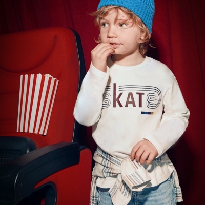 Criança com gorro azul e camisola branca com texto SKAT ao lado de cadeira vermelha e pipoca