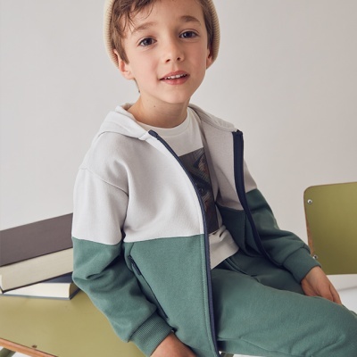 Menino com casaco branco e verde sentado numa cadeira verde junto a secretária de madeira.