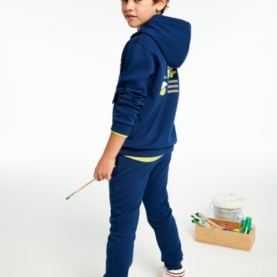 Menino com fatos de treino azuis segurando pincel perto de caixa de pinceis e balde