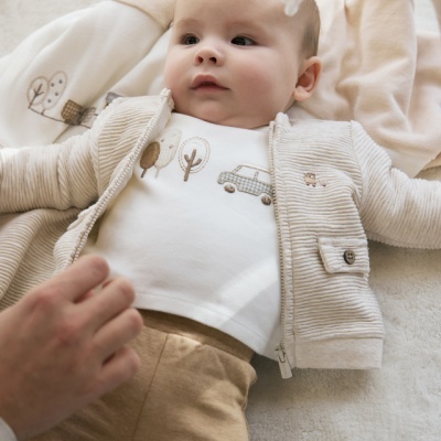 Bebé deitado com roupa bege e branca decorada com árvores e carro, sobre superfície branca com mó de penas