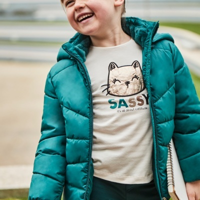 Menina com casaco verde-petróleo e camisola creme com desenho de gato sorrindo ao ar livre