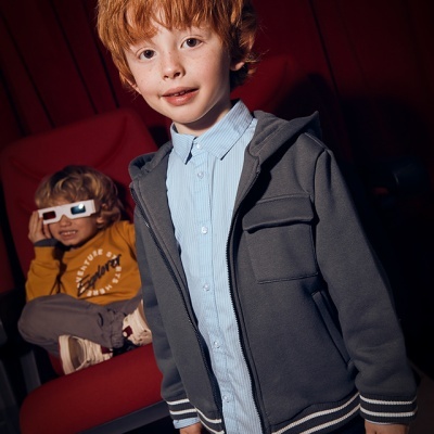 Criança com casaco cinzento e camisa azul em cinema com cadeiras vermelhas e criança ao fundo com óculos 3D