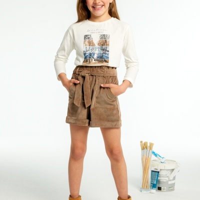 Menina com camisola branca, calções castanhos e botas castanhas perto de balde de tinta com pincéis