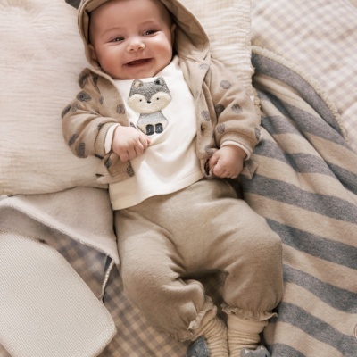 Bebé sorridente vestido com casaco bege com orelhas, camisola com raposa, calças e meias com rosto de animal, em cama com lençóis xadrez cinza e branco