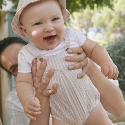 Bebé com chapéu e jardineira às riscas segurado por adulto ao ar livre