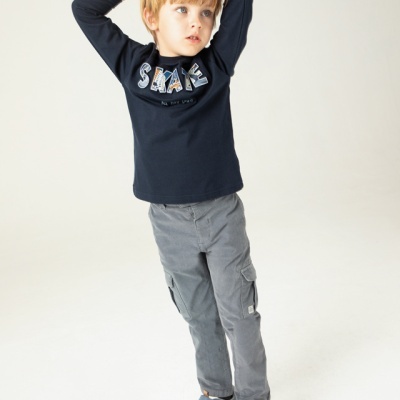 Menino com t-shirt azul escuro, calças cinza e sapatilhas azuis em fundo branco