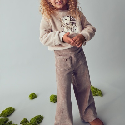 Criança com roupa bege felpuda e sapatos marrons em fundo cinza com musgo verde