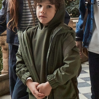 Criança com casaco verde com capuz e calças jeans azuis em ambiente exterior com chão xadrez.