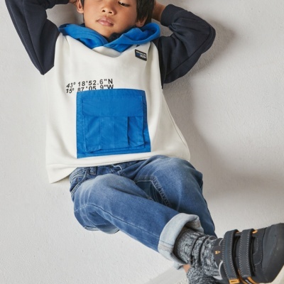 Menino com casaco branco e azul, calças de ganga azuis e sapatilhas pretas sentado contra parede branca