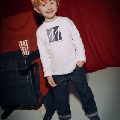 Criança com camisola branca e estampado geométrico, calças escuras e sapatos cinzentos ao lado de cadeira preta e pacote de pipocas listrado