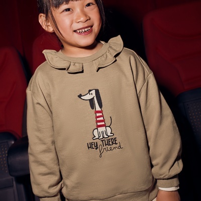 Camisola bege com estampa de cão e texto, usada por criança em ambiente com cadeiras vermelhas