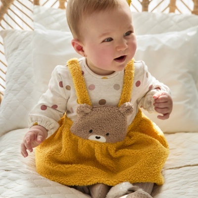 Bebé com vestido amarelo e ursinho sentado em cama com cabeceira em vime