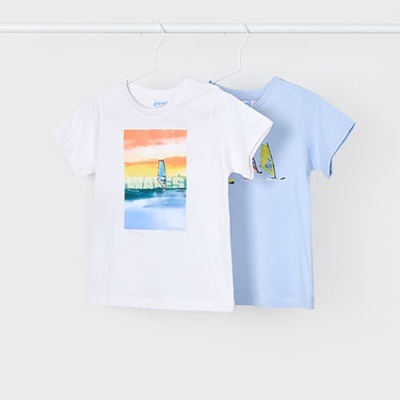 Dois tshirts de criança pendurados, uma branca com estampa de vela sobre mar colorido e outra azul clara com estampa de vela