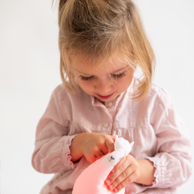 Luz LED de presença em forma de gato emitindo luz rosa nas mãos de uma criança com vestido rosa claro.