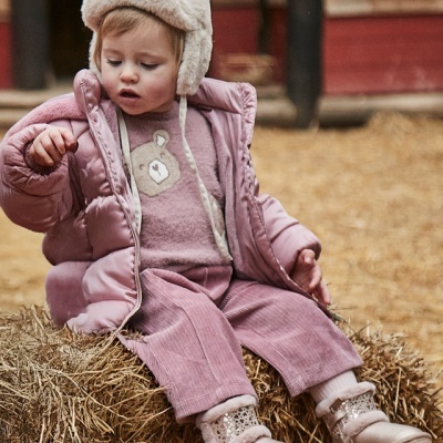 Criança sentada em fardo de palha com roupa rosa e gorro bege com orelhas