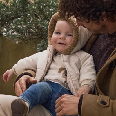 Bebé com roupa clara sentado no colo de adulto com casaco castanho
