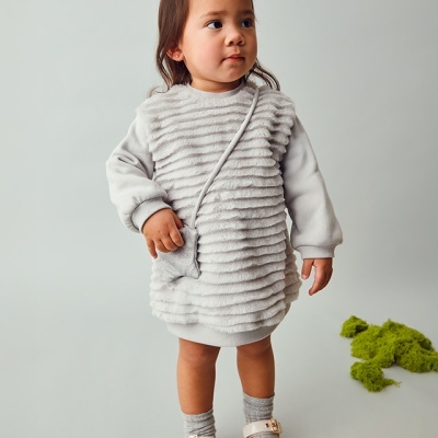 Menina com vestido cinza texturizado, meias cinzentas e sapatos brancos, sobre fundo cinzento com almofadas verdes no chão.