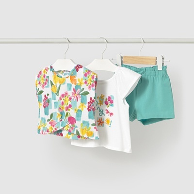 Conjunto infantil de três peças com blusa floral, t-shirt branca estampada e calções verdes, pendurado numa barra branca.