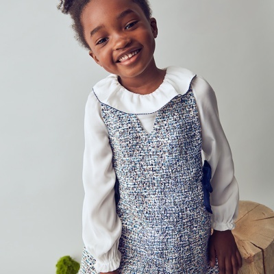 Menina sorridente com vestido tipo tweed azul e blusa branca com gola rodada e folhos