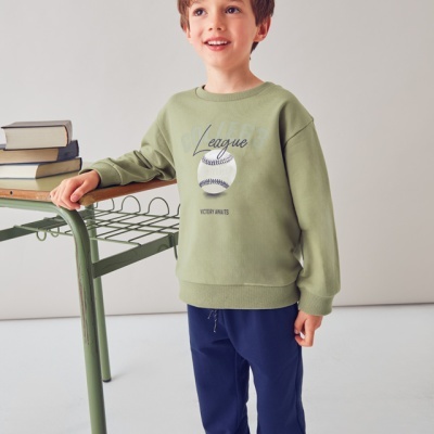 Criança com camisola verde de basebol e calças azuis junto a mesa verde com livros