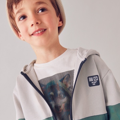 Criança a usar casaco com capuz verde e bege e camisola branca com estampa