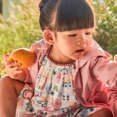 Criança com vestido floral e casaco rosa segurando tangerina ao ar livre