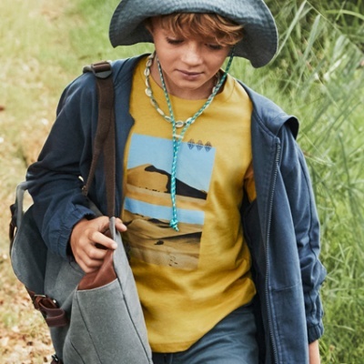 Rapaz criança com camisola amarela e casaco azul a caminhar com mochila e chapéu cinza ao ar livre