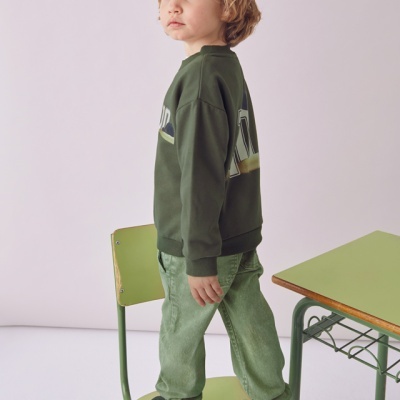 Criança com sweat verde escuro e calças verdes em cima de cadeira verde junto a secretária verde.