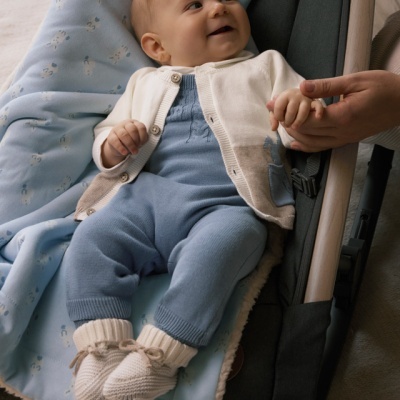 Bebé vestido com conjunto azul e cardigan branco deitado em suporte cinzento com cobertor azul claro com flores brancas
