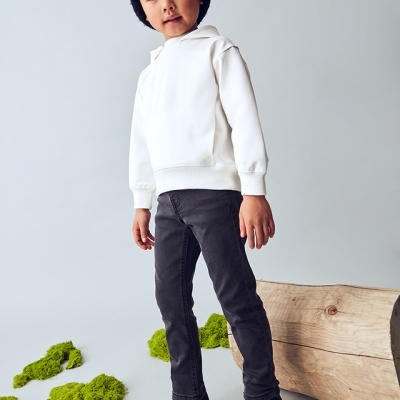 Criança com hoodie branco, calças e botas pretas em estúdio com tronco de árvore e musgo.