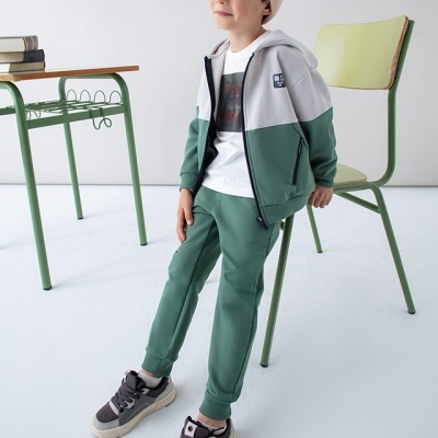 Criança com roupa desportiva verde e branca e sapatilhas cinzentas, sentada numa cadeira verde ao lado de uma mesa com livros.